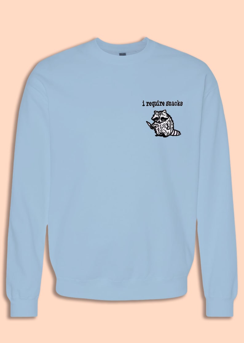 May include: Light blue sweatshirt featuring a graphic of a raccoon holding a knife, with the text "i require snacks" above the raccoon. The raccoon is black and white with a pink and white striped tail. The sweatshirt is a crew neck.