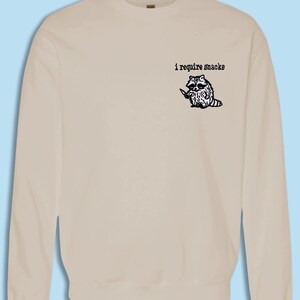 May include: Beige sweatshirt with a graphic of a raccoon holding a knife, accompanied by the text "i require snacks" in black. The raccoon is black and white with a purple tail. The sweatshirt is a solid color.