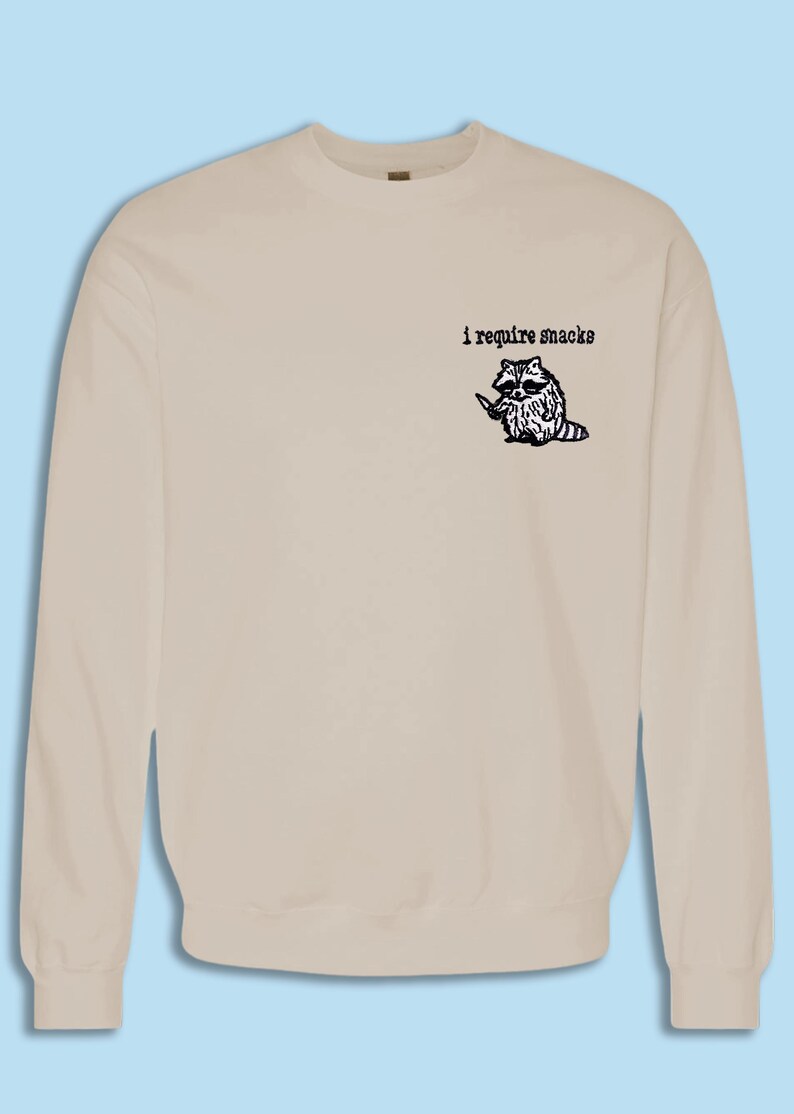 May include: Beige sweatshirt with a graphic of a raccoon holding a knife, accompanied by the text "i require snacks" in black. The raccoon is black and white with a purple tail. The sweatshirt is a solid color.