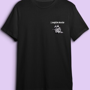 May include: Black t-shirt with a white graphic of a raccoon holding a knife, and the text "i require snacks". The raccoon illustration is detailed and the text is in a simple font.