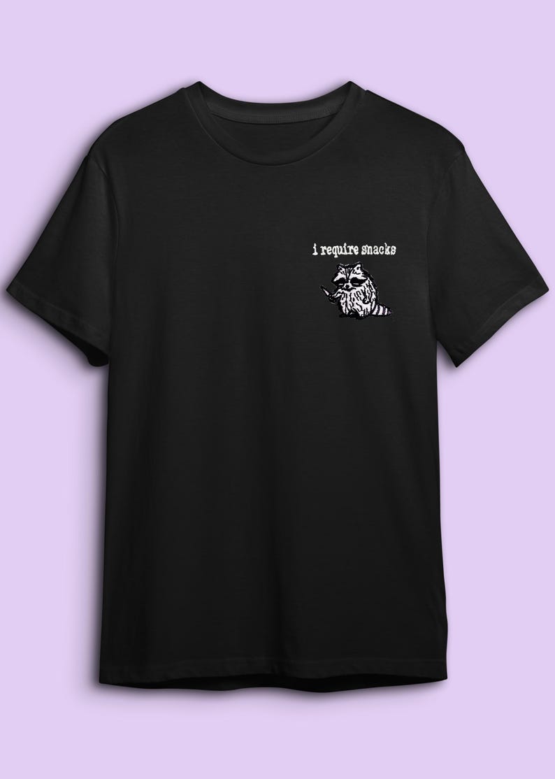 May include: Black t-shirt with a white graphic of a raccoon holding a knife, and the text "i require snacks". The raccoon illustration is detailed and the text is in a simple font.