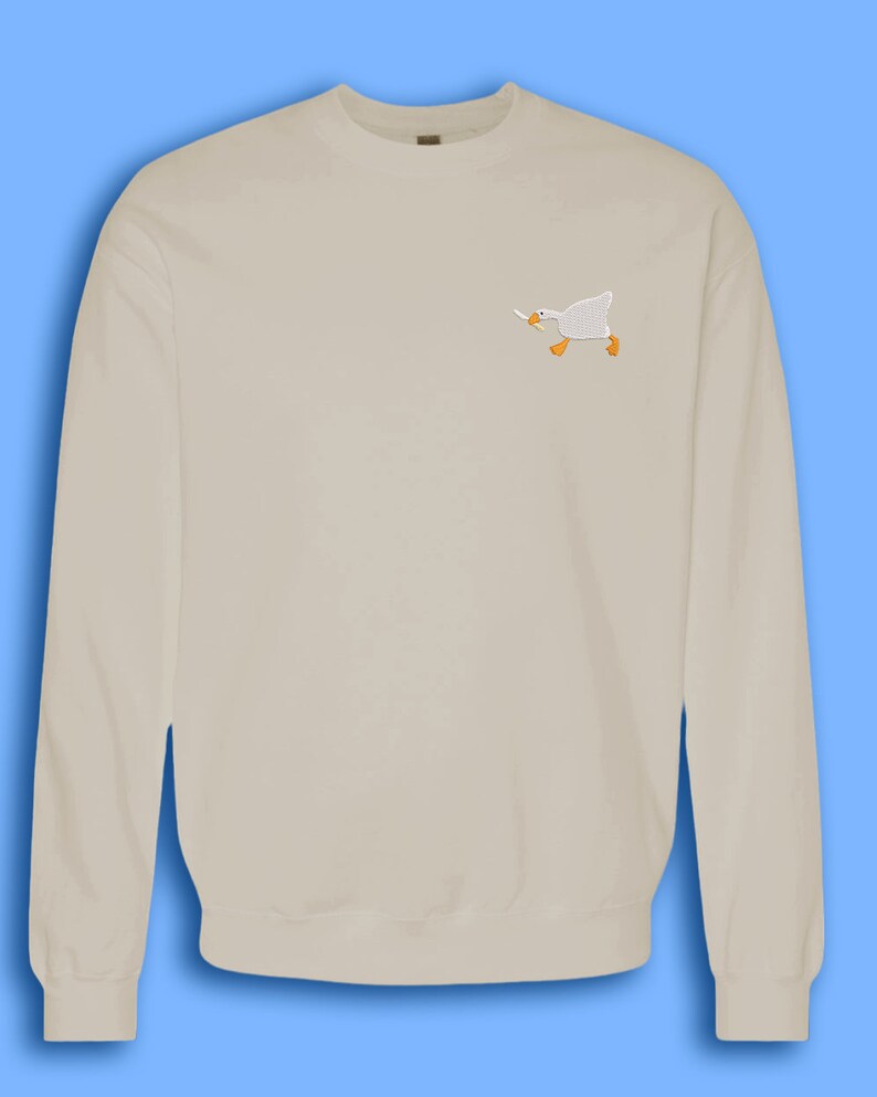 May include: A light beige sweatshirt with a small embroidered image of a white goose holding a carrot in its beak.