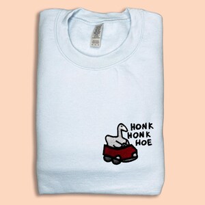 A white t-shirt with a small embroidered image of a cartoon goose sitting in a red car, along with the text "HONK HONK HOE" printed on it.