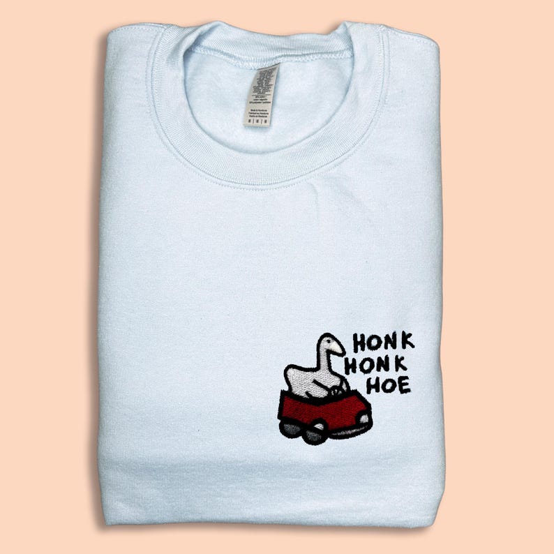 A white t-shirt with a small embroidered image of a cartoon goose sitting in a red car, along with the text "HONK HONK HOE" printed on it.