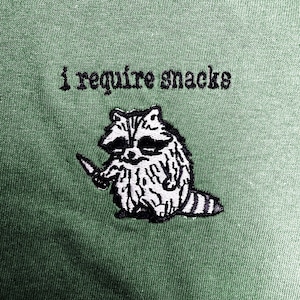 A raccoon holding a knife and the text "I require snacks" is embroidered on a green fabric.