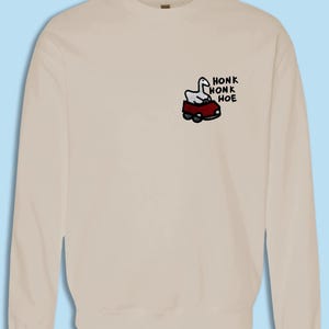 A beige sweater with a small cartoon image of a car and the text "HONK HONK HOE" printed on it.