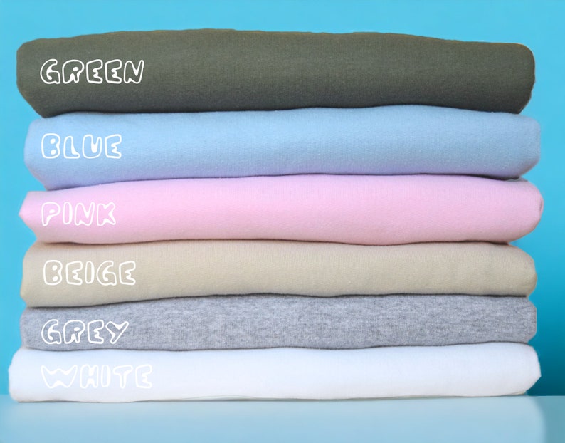 May include: A stack of six folded t-shirts in different colors: green, blue, pink, beige, gray, and white. The colors are labeled in white text.