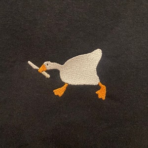 May include: A white goose with orange feet holding a knife in its beak, embroidered on a black t-shirt.