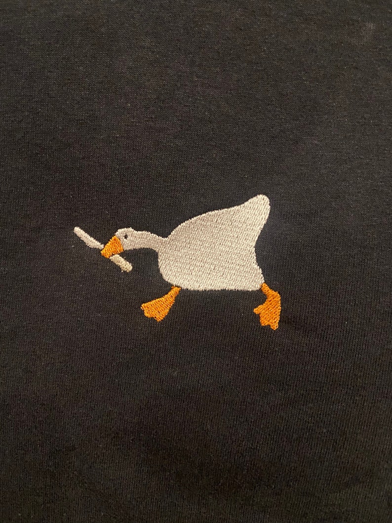 May include: A white goose with orange feet holding a knife in its beak, embroidered on a black t-shirt.