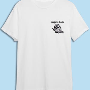May include: White t-shirt featuring a black illustration of a raccoon holding a knife, with the text "i require snacks" above it. The shirt is made of a soft material and has short sleeves.