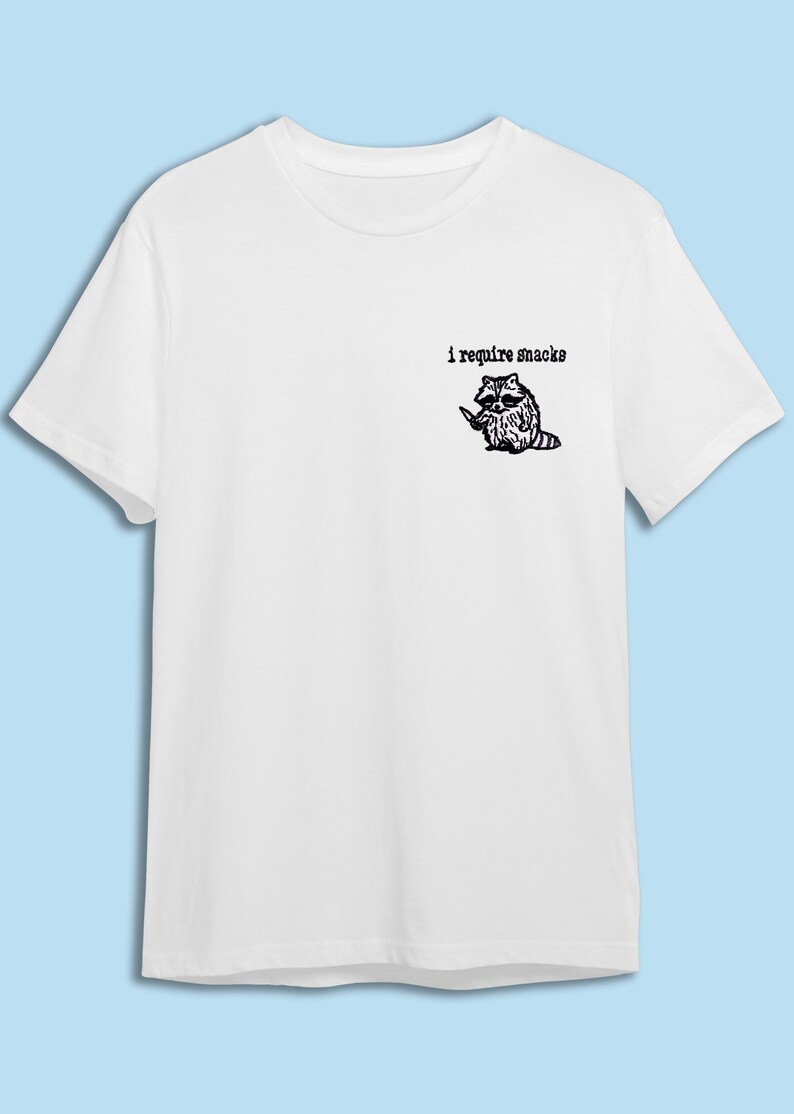 May include: White t-shirt featuring a black illustration of a raccoon holding a knife, with the text "i require snacks" above it. The shirt is made of a soft material and has short sleeves.
