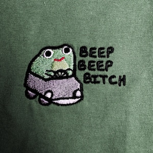 a green frog with a car, accompanied by the text "BEEP BEEP BITCH" in a playful, humorous manner.