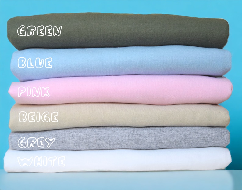 May include: A stack of six folded t-shirts in different colors: green, blue, pink, beige, grey, and white. The colors are labeled in white text.