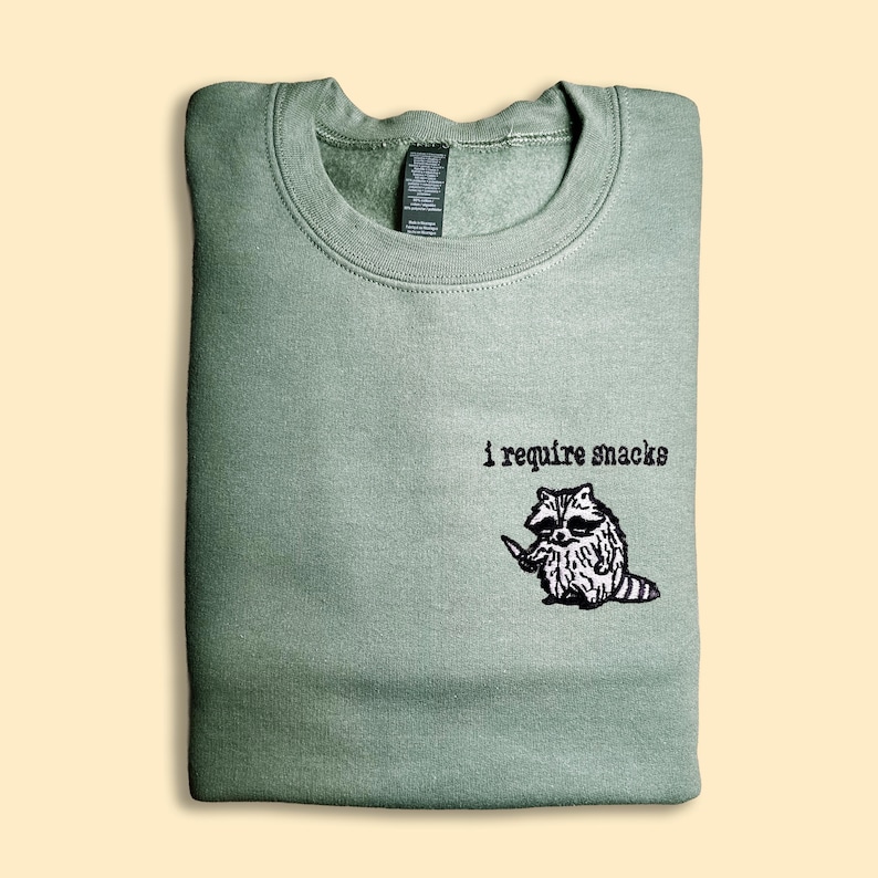 A green sweatshirt with a black and white raccoon graphic and the text "I require snacks" printed on it.