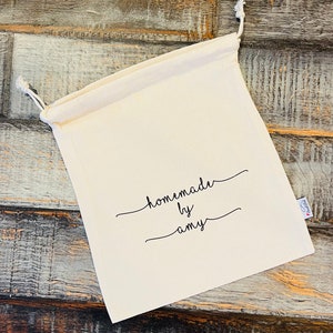 May include: Cream-colored drawstring bag with the text "homemade by amy" in black script. The bag is made of a soft fabric and has a white drawstring closure. A small tag is attached to the side.