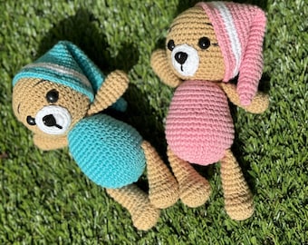 May include: Two crocheted teddy bears wearing sleep caps. One bear is wearing a light blue sleep cap and a light blue outfit. The other bear is wearing a pink sleep cap and a pink outfit. The bears are lying on a green grassy surface.