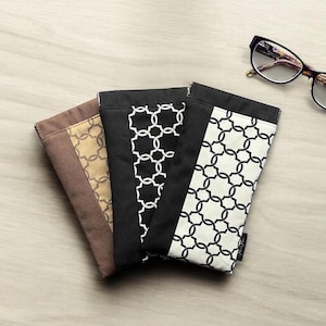 Puede incluir: Tres estuches para gafas con estampado en negro, marrón y blanco con un diseño geométrico. Los estuches están doblados juntos sobre una superficie de madera clara. Un par de gafas está sobre la superficie a la derecha de los estuches.