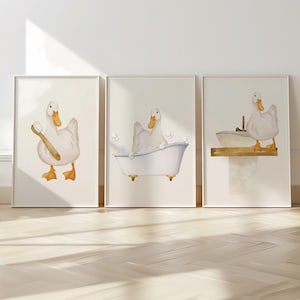 May include: Three framed prints featuring a white duck in different scenes. The first print shows the duck holding a toothbrush, the second shows the duck in a bathtub, and the third shows the duck standing on a ledge.