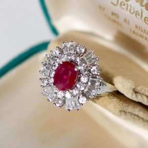 Puede incluir: Un anillo de rubí con un racimo de diamantes engastados en una banda de metal blanco. El anillo está en una caja de anillo de terciopelo verde con las palabras "Mei Yu Jewellery Co." impresas en el interior.