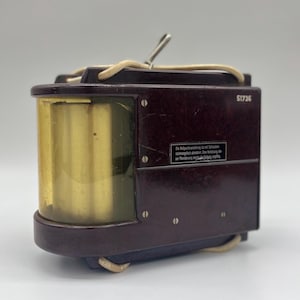 May include: A dark brown vintage electrical component with a clear glass window and a label that reads "51736.  Zu  Abgleichverstimmung  ist  der  Kondensator  mit  einem  Schraubendreher  an  der  Unterseite  des  Gehäuses  zu  verstellen.  Die  Betriebsspannung  ist  auf  dem  Typenschild  angegeben."