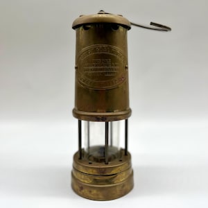 May include: Vintage brass miner's lamp with a clear glass cylinder and a handle. The lamp has the text "BRITISH COAL MINING COMPANY" and other text. The lamp is a cylindrical shape and has a hook on top.