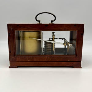 May include: A vintage wooden box with a glass front and a brass mechanism inside. The box has a handle on top and sits on a white surface.