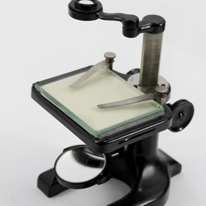 May include: A vintage black microscope with a silver metal column and a small mirror. The microscope has a rectangular stage with a glass slide and two metal clips. The eyepiece is black and round.