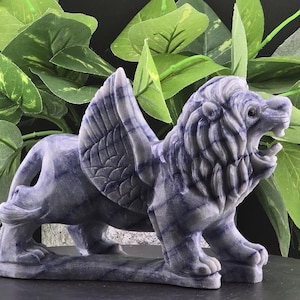 May include: A carved stone sculpture of a winged lion, featuring a detailed mane and wings. The lion is in a roaring pose, with a blue and white color scheme. The sculpture is set against a backdrop of green foliage.