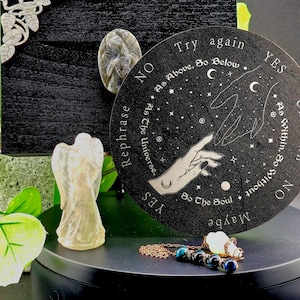 May include: A black circular pendulum board with a silver hand and the words "Try again", "As Above, So Below", "Yes", "As The Universe, Rephrase", "So The Soul", "Maybe", "As Within, So Without", "No", and "Yes" inscribed around the edge. The board is sitting on a black surface with a small angel figurine and a copper chain with blue and black beads.