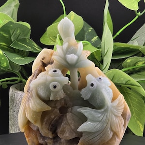 May include: A jade carving of two fish swimming around a lotus flower. The carving is a light green color with brown accents.