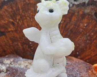 May include: White jade dragon figurine with green accents. The dragon is sitting with its wings spread out and its head tilted to the side. It has black eyes and a small, pointed snout.