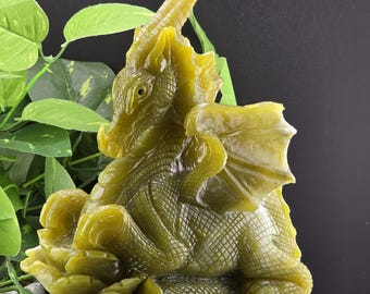 Puede incluir: Una detallada escultura de dragón verde con escamas texturizadas, alas y una cara detallada. El dragón está posado sobre una base de madera, con follaje verde en el fondo. Es un objeto decorativo.