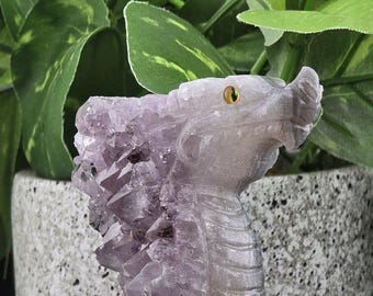 May include: A purple amethyst dragon head sculpture with a detailed design and a single yellow eye. The sculpture is carved from a natural amethyst crystal.