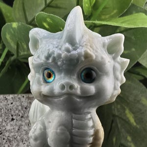 May include: A small, light blue dragon figurine carved from jade with blue eyes. The dragon is sitting on its hind legs with its wings folded behind it.