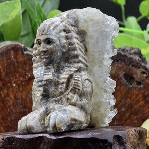 May include: A carved stone sphinx sculpture with a quartz crystal attached to its back. The sphinx is grey and tan, with detailed facial features and a regal pose. The sculpture is on a dark brown base, with the text "The Mystical Manor".