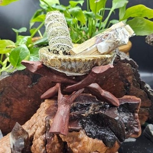 May include: A wooden stand with a bowl on top, holding two bundles of sage smudge sticks. The stand is on a piece of wood with a brown and black surface.