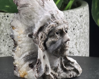 May include: A white and gray carved stone sculpture of a Native American chief wearing a headdress. The sculpture is detailed and realistic, with a feathered headdress and a stern expression.