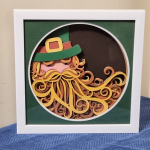 May include: A paper craft of a leprechaun with a green hat and a curly orange beard. The leprechaun is framed in a white frame with a green mat.