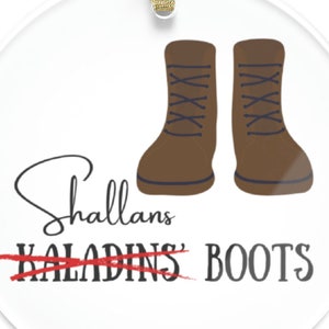 May include: A round, white, glass ornament with a gold ribbon. The ornament features a pair of brown boots with laces and the text "Shallans' Kaladins' Boots" with a red line through "Kaladins'"