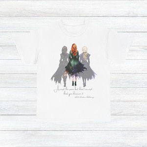 Può includere: T-shirt bianca con un disegno ad acquerello di tre donne con mantelli fluenti. La figura centrale ha i capelli rossi e un vestito verde. Il testo recita: "Accetta il dolore ma non accettare che lo meriti."