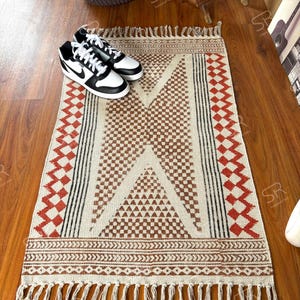 Peut inclure: Un tapis rectangulaire crème avec un motif géométrique marron, noir et rouge. Le tapis présente un motif central en losange et des bords à franges. Une paire de baskets noires et blanches est posée sur le tapis.