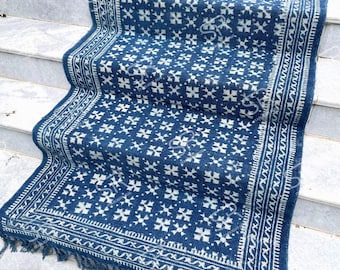 Op de afbeelding: Een blauw en wit patroonkleed met een geometrisch ontwerp. Het kleed heeft een donkerblauwe achtergrond met witte bloemachtige vormen en een witte rand. Het kleed is rechthoekig met franjes aan de uiteinden.