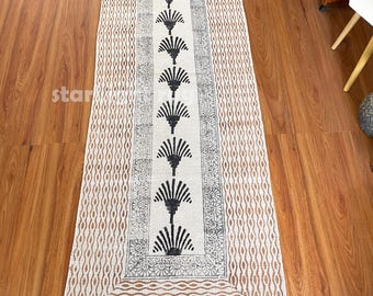 Op de afbeelding: Een rechthoekig beige en wit patroonkleed met een centraal ontwerp van zwarte waaiervormige vormen. Het kleed heeft een gefranjerde rand en ligt op een houten vloer. Het kleed is ongeveer 152 cm lang.