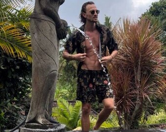 Puede incluir: Una persona con una camisa negra con estampado floral y pantalones cortos a juego está de pie en una piscina. La persona lleva gafas de sol y sostiene una cadena de agua. A la izquierda hay una estatua de piedra.