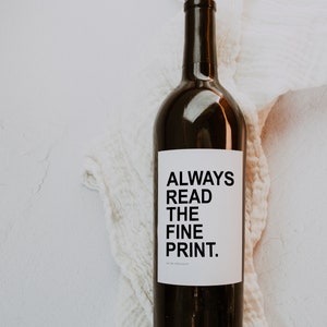May include: A dark brown wine bottle with a white label that reads "ALWAYS READ THE FINE PRINT." The bottle is set against a white, textured background.