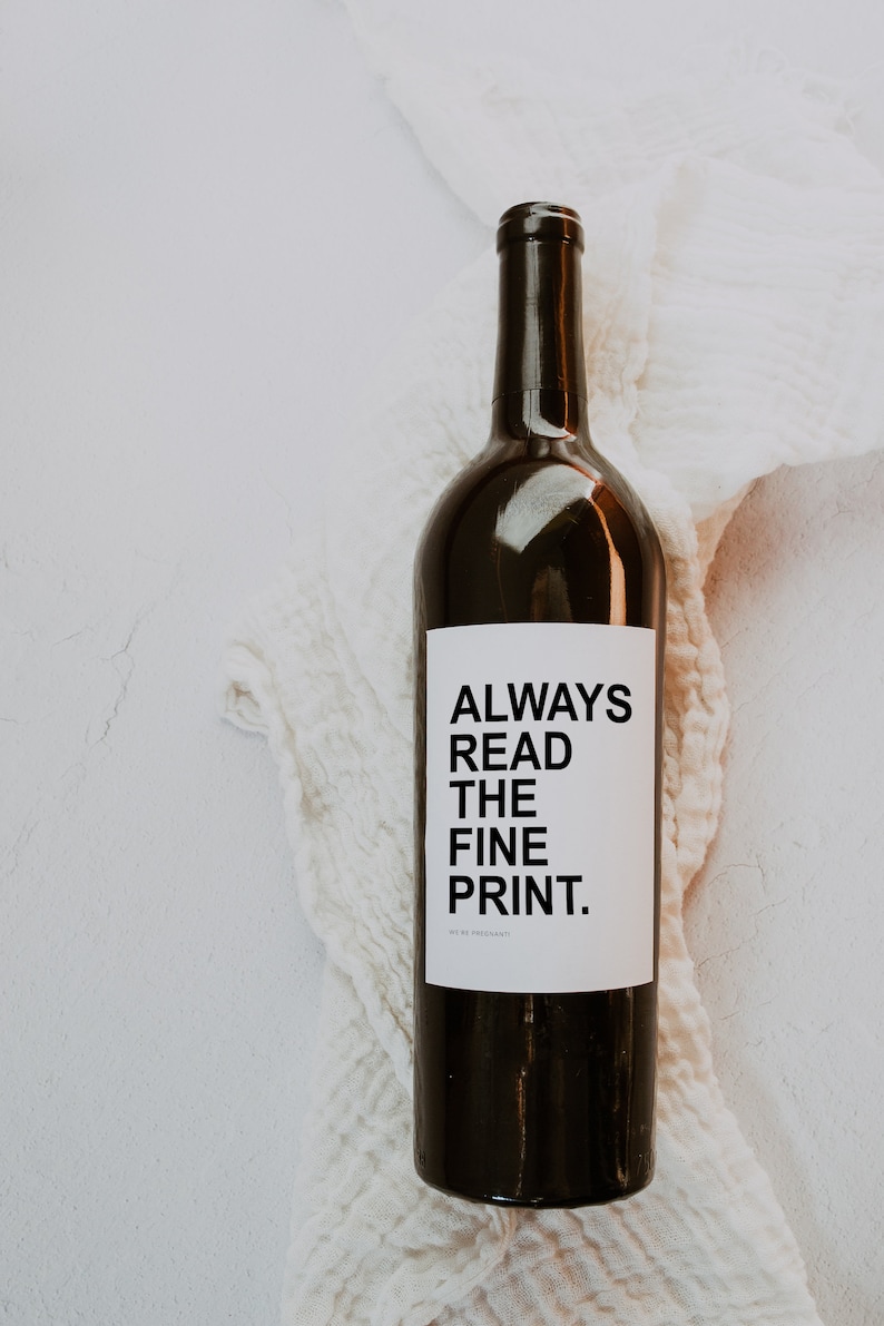 May include: A dark brown wine bottle with a white label that reads "ALWAYS READ THE FINE PRINT." The bottle is set against a white, textured background.