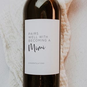 May include: A white wine bottle label with black text that reads "Pairs well with becoming a Mimi. Congratulations!"