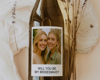 Op de afbeelding: Een bruine wijnfles met een etiket met twee lachende vrouwen. Op het etiket staat "WILL YOU BE MY BRIDESMAID?" Gedroogde lavendel takken zijn naast de fles geplaatst. De achtergrond is een zachte, neutrale kleur.