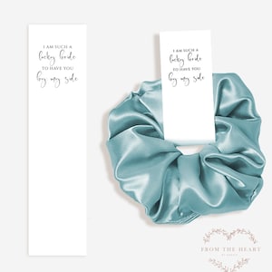 May include: A light blue satin scrunchie with a white card attached to it. The card has the text "I am such a lucky bride to have you by my side" written on it. The card is from "From the Heart by 300sie".