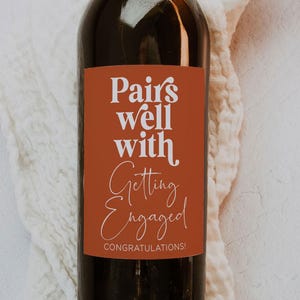 Op de afbeelding: Een bruine wijnfles met een etiket waarop staat "Pairs well with Getting Engaged! Congratulations!" Het etiket is oranje-bruin van kleur met witte tekst.